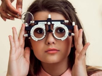 horizontal-shot-interested-curious-caucasian-girl-appointment-with-eye-care-specialist-wearing-phoropter-while-ophthalmologist-checking-her-vision-sitting-yellow-wall